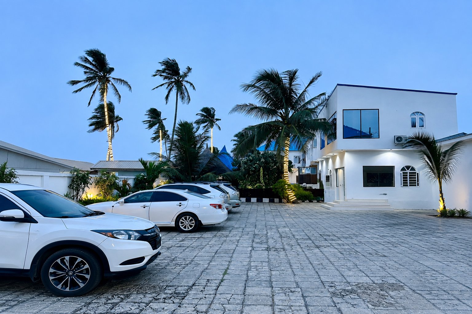 Jet Beach & Hotel: palm trees and guest parking court beside the white two-storey hotel building at dusk
